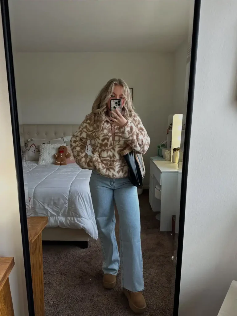Petite blonde woman taking a mirror selfie in a cozy bedroom decorated with holiday pillows and a gingerbread plush toy on the bed, wearing a cream and warm tan botanical-print sherpa teddy fleece zip-front jacket, light wash wide-leg jeans with a raw frayed hem at the ankle, and tan UGG-style short platform boots, holding a black bag tucked under one arm, with a white vanity desk and lamp visible to the right.
