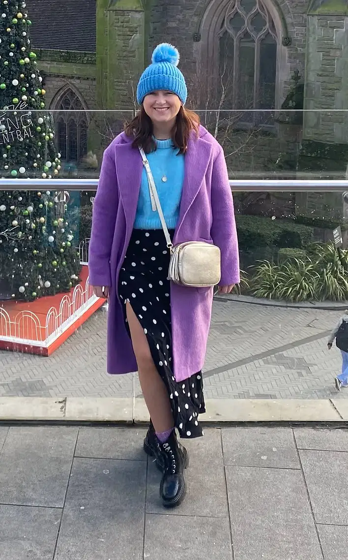 Purple Coat with Blue Knit and Polka Dot Midi Skirt, petite woman in open purple coat, blue turtleneck knit, black polka dot slit midi skirt and black combat boots with blue pom beanie outdoors.
