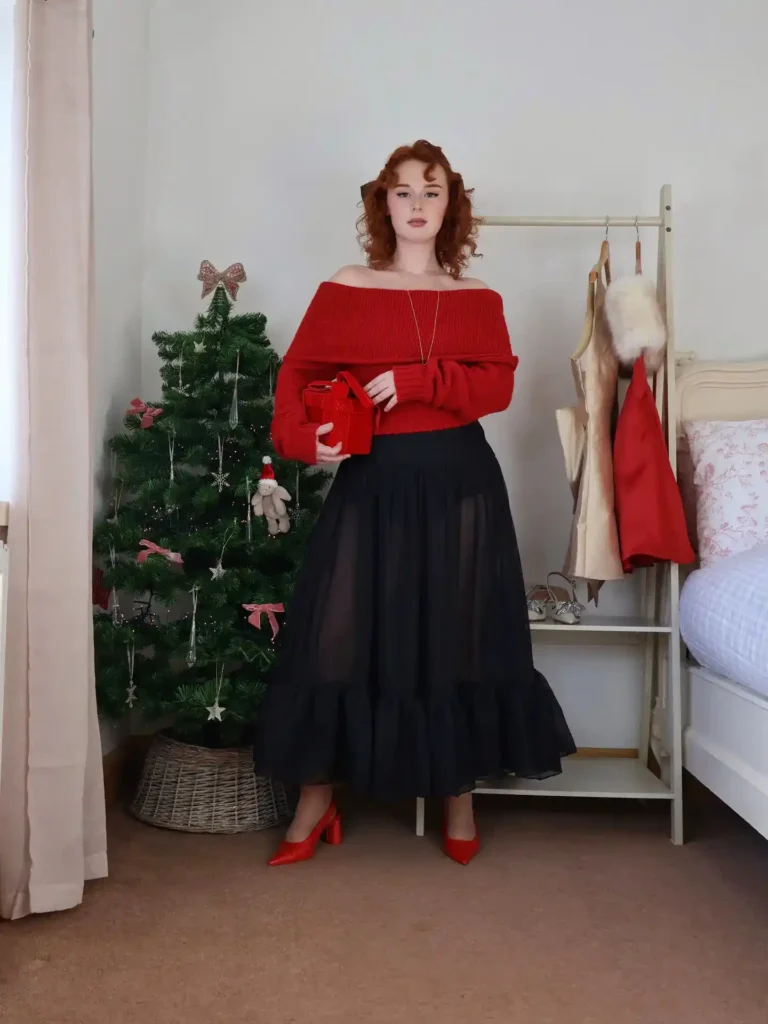 Petite woman in a red chunky off-shoulder bardot knit sweater with wide draped neckline and balloon sleeves, paired with a floor-length black sheer chiffon tiered ruffle maxi skirt, wearing red patent leather pointed-toe block heel pumps, holding a small red velvet structured clutch, with a long delicate gold pendant necklace and a dark bow hair clip in her auburn curly hair, standing beside a small decorated Christmas tree in a wicker basket with pink ribbon bows and silver star ornaments in a softly lit bedroom.