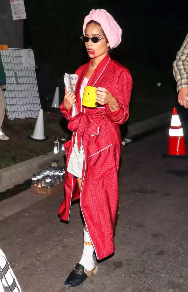Woman wearing a red satin polka dot dressing robe with white piping trim and belt tied at the waist over a white striped pajama set, accessorized with a pink terrycloth towel wrapped on the head and small dark cat-eye sunglasses, with dripping red Halloween makeup at the mouth, holding a yellow smiley face mug and a folded newspaper, wearing white knee-high socks and black loafer mules, photographed outdoors at night.