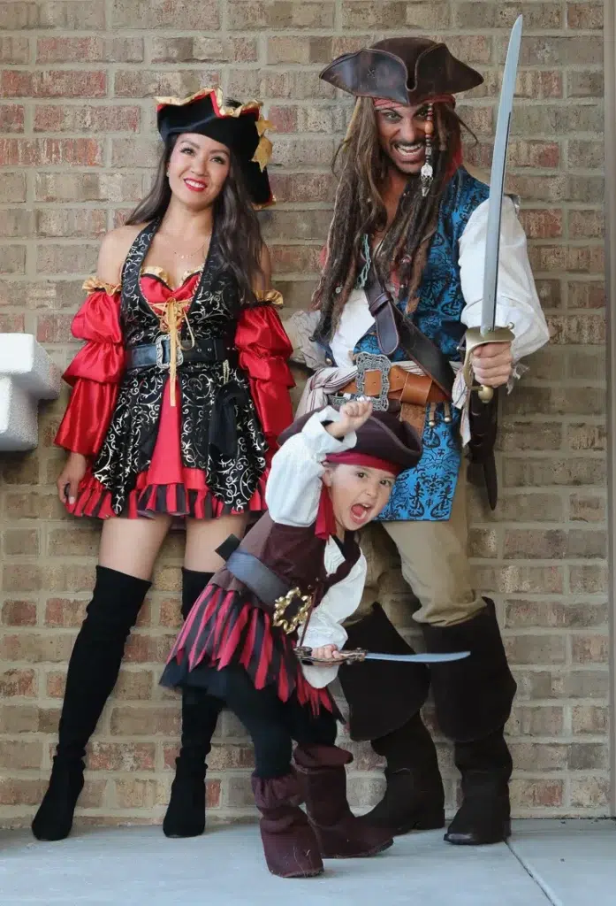 Petite woman with long dark wavy hair and red lips wearing a red and black ruffled pirate costume mini dress with a corset-style bodice featuring gold scroll detailing, red satin puff sleeves, and layered ruffle skirt, paired with tall black over-the-knee boots and a black pirate hat with gold trim accent, posing and smiling against a brick wall.