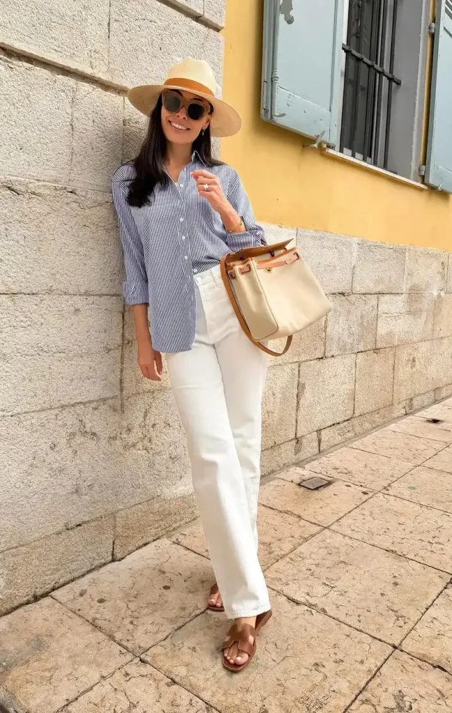 Petite woman with long dark hair wearing white relaxed straight-leg cropped denim jeans, an oversized blue and white fine vertical-stripe button-down shirt worn untucked at hip length, a wide-brim natural straw panama hat, round brown-tinted sunglasses, tan leather flat slide sandals, and carrying a cream canvas bucket bag with tan leather trim and a wooden top handle, leaning against a stone block wall on a European street with a yellow-painted building facade and blue-shuttered window visible in the background.