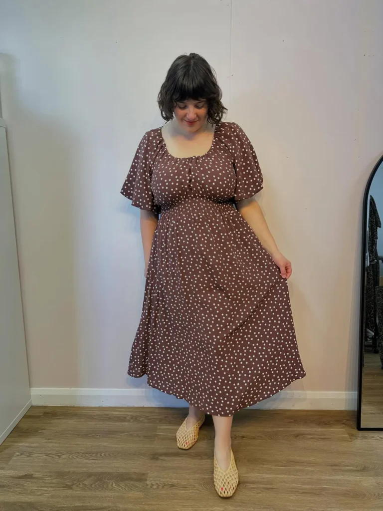 Petite plus-size woman with long dark wavy hair and medium gold teardrop hoop earrings, smiling and walking forward on a tree-lined city sidewalk, wearing a rich rust terracotta crinkle-texture short flutter-sleeve midi dress with a sweetheart neckline, a front tie-bow gathering at the center bust, a fitted gathered waistline, a centre-front button placket running from the waist to a softly flounced midi hem, carrying a round woven crochet tote bag with tan leather handles, and wearing cognac brown wedge sandals with ankle straps, accessorised with layered gold chain necklaces, gold bracelets, and a gold watch.