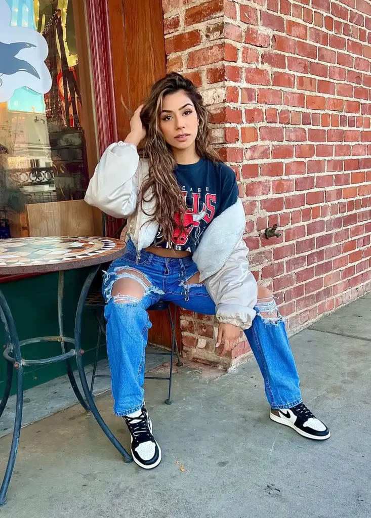 Petite woman seated outside a brick-front boutique wearing bright medium-wash heavily ripped boyfriend jeans, a cropped navy Chicago Bulls graphic tee layered under an open cream satin bomber jacket with contrast sleeves, holding a soft cream cloud-style bag, with black and white high-top sneakers.