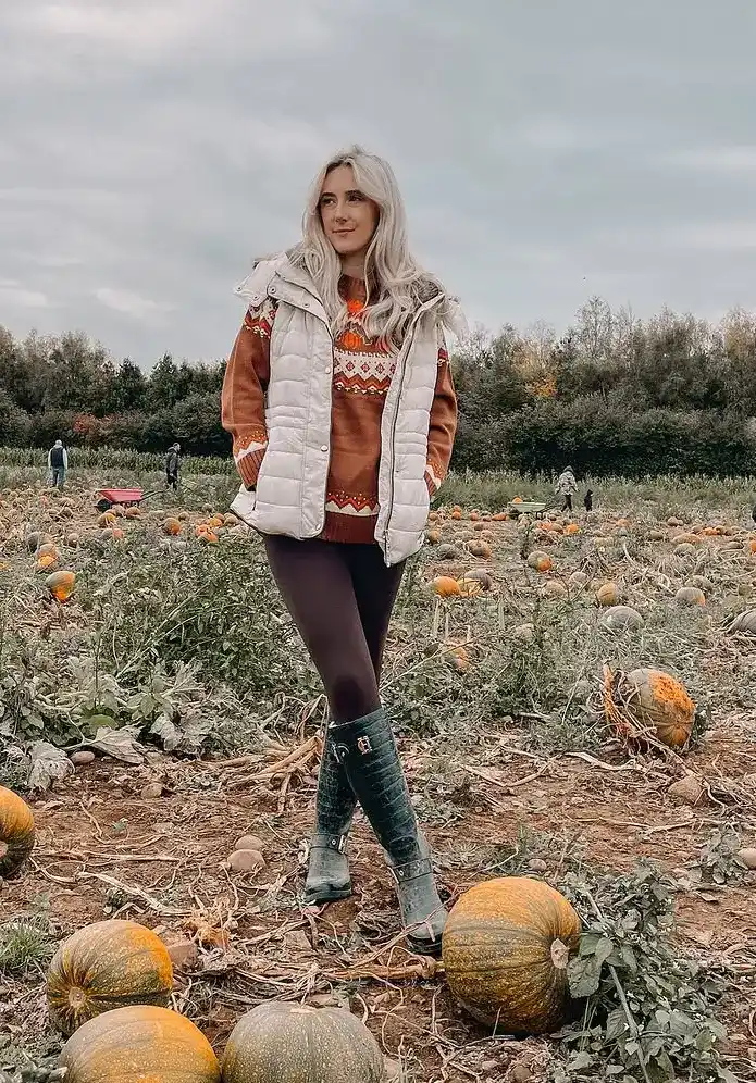 Petite woman in a rust and cream Fair Isle yoke-pattern knit sweater layered under an open cream quilted puffer vest, with dark chocolate brown opaque leggings and dark green rubber wellington boots with buckle detail, standing confidently in a wide open pumpkin field on a grey overcast day with large green and orange pumpkins on the ground around her.