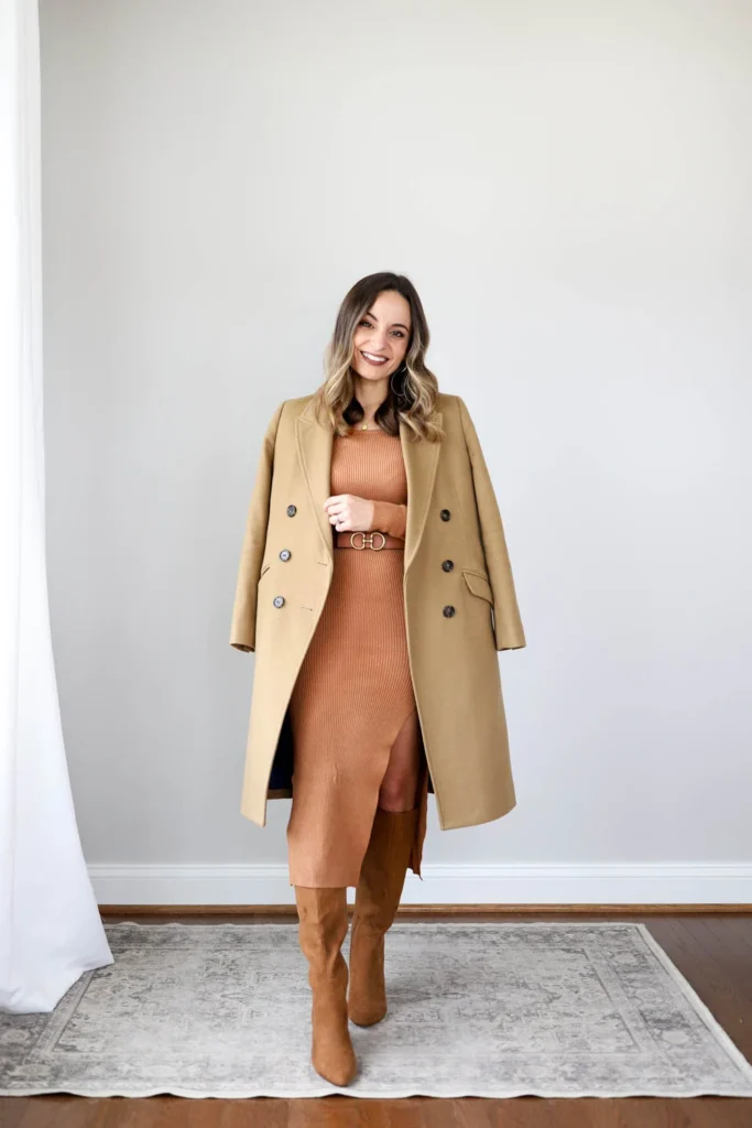 Petite woman with wavy highlighted brown hair standing in a neutral indoor room on a vintage-style area rug, wearing a camel longline double-breasted coat worn open, a rust terracotta fitted ribbed knit midi dress with a front slit, a wide tan belt with a gold double-ring buckle cinched at the natural waist over the dress, tall camel suede knee-high boots, gold hoop earrings, and a delicate gold layered necklace, smiling with one hand holding the coat lapel.
