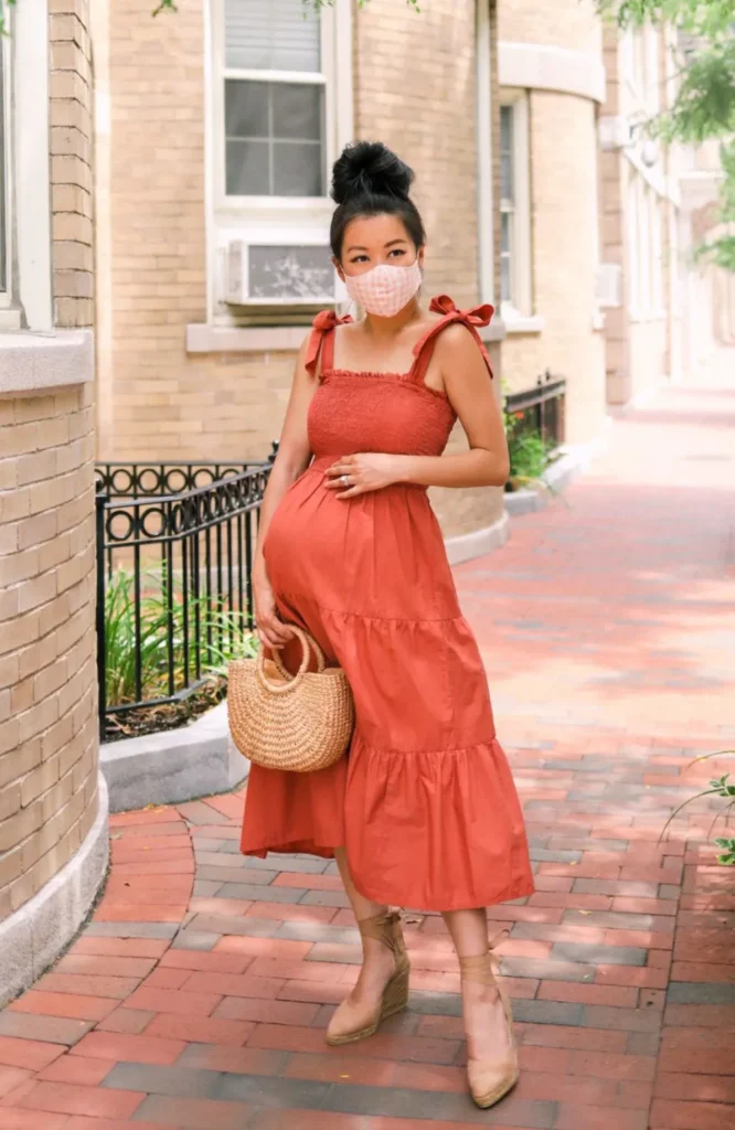 Petite pregnant Asian woman with dark hair in a high bun, wearing a solid rust terracotta smocked-bodice tiered sundress with adjustable tie-bow shoulder straps, carrying a small round woven rattan circle tote bag by its top handle, wearing camel espadrille wedge sandals with wraparound ankle lace ties and a light pink gingham face mask, standing on a red brick city sidewalk in front of warm sandy brick brownstone buildings with an iron fence railing and green trees in the background.