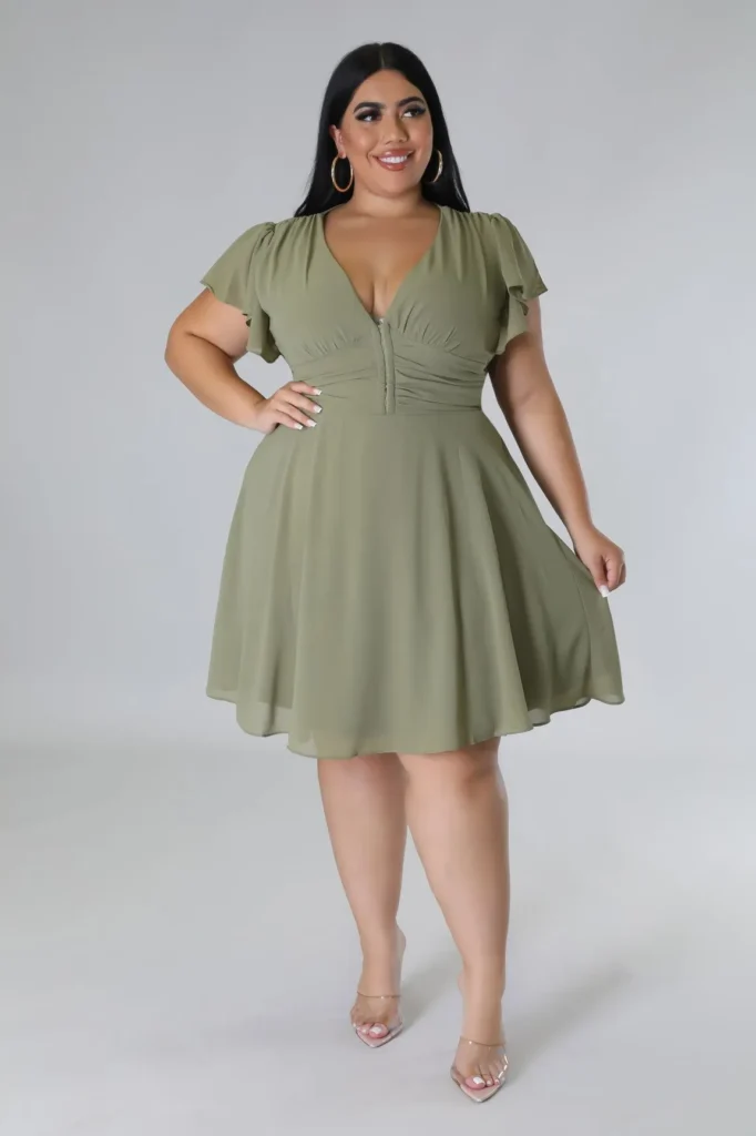 Petite plus-size woman with long straight dark hair, wearing bold gold hoop earrings and smiling while looking slightly to the side with one hand on her hip, dressed in a sage olive green chiffon fit-and-flare mini dress with a deep V-neckline, short flutter sleeves, diagonal ruching across the bust radiating from a center-front hook closure, horizontal ruching across the fitted waistband, and a full A-line circle skirt landing above the knee, paired with clear perspex strappy heeled sandals and light-coloured manicured nails, photographed against a plain neutral grey studio backdrop.
