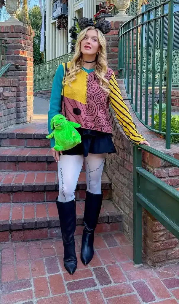 Petite woman with long blonde wavy hair wearing a Sally from Nightmare Before Christmas inspired Halloween outfit consisting of a multicolor patchwork knit sweater with teal, mustard yellow, pink swirl, and black striped panels, a black satin mini skirt, white thigh-high stockings with black stitching detail, and tall black over-the-knee heeled boots, accessorized with a black velvet Minnie Mouse ear headband and a black choker necklace, holding a green character plush, standing on brick stairs outside a Halloween-decorated Victorian building.