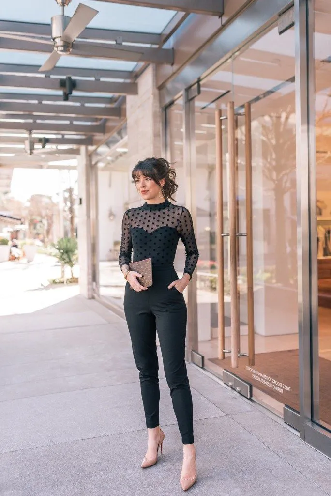 Petite woman in a black sheer polka dot mesh long-sleeve mock-neck bodysuit tucked into high-waisted fitted black cigarette trousers with a tapered ankle length, wearing nude pointed-toe stiletto heels and holding a tan monogram print envelope clutch, with gold hoop earrings and hair styled in a loose updo with fringe bangs, standing outside a modern glass-fronted building.