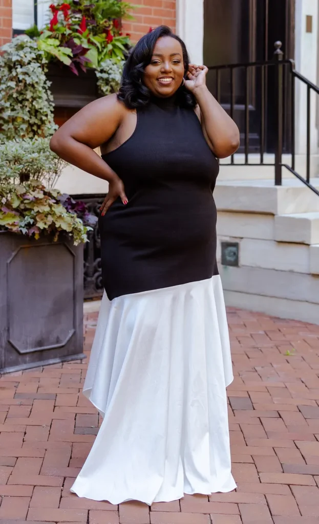 Petite plus-size woman with wavy shoulder-length dark hair and small gold hoop earrings, smiling with one hand raised near her face, wearing a two-tone colorblock maxi dress with a fitted black ribbed sleeveless halter neckline bodice and a flowing white bias-cut satin maxi skirt, standing on brick pavers outside an urban building with lush flowering plants and wrought iron railings in the background.