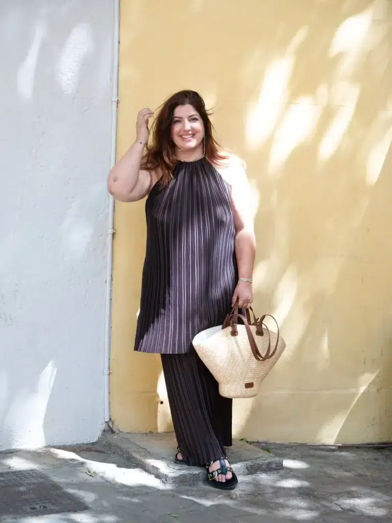 Petite plus-size woman with long loose auburn-brown hair, smiling and raising one hand to her hair, wearing a matching dark charcoal pleated satin sleeveless halter tunic top with a gathered neckline and coordinating wide-leg pleated satin pants creating a full-length monochromatic column silhouette, carrying a large natural straw woven basket tote bag with tan leather top handles, wearing black strappy flat sandals with metal hardware detail, styled with a delicate chain necklace and thin stacked bracelets, standing against a sunny yellow exterior wall with dappled tree shadow.