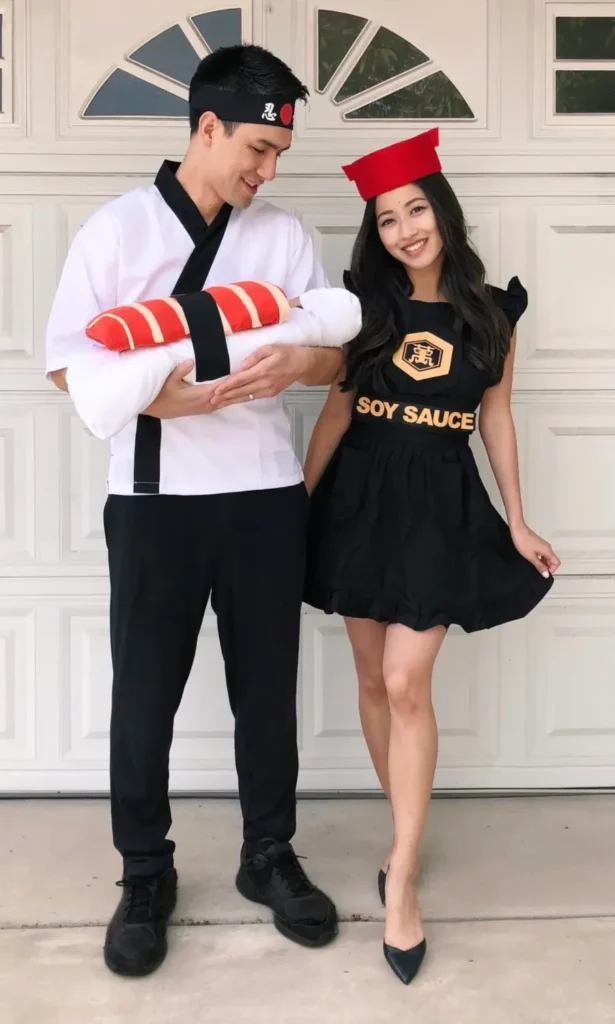 Petite woman with long dark wavy hair wearing a black Halloween soy sauce bottle costume mini dress with flutter ruffle sleeves, a gold hexagonal logo and "Soy Sauce" text printed on the fitted bodice, and a slightly bubble-shaped mini skirt, paired with dark pointed-toe heels and a small red cylindrical cap on the head, posing with a male partner in a sushi chef costume outside a residential garage.