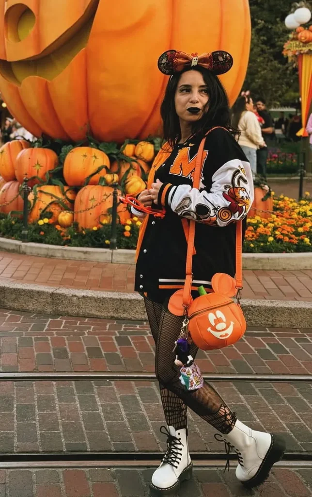 Petite woman with dark hair and black lipstick wearing a black and orange Halloween varsity letterman jacket with character graphics on the sleeves, black fishnet tights, and white lace-up combat boots, accessorized with a Halloween sequin Minnie Mouse ear headband and an orange pumpkin-shaped character crossbody bag, posing in front of large seasonal pumpkin park displays and orange floral arrangements.