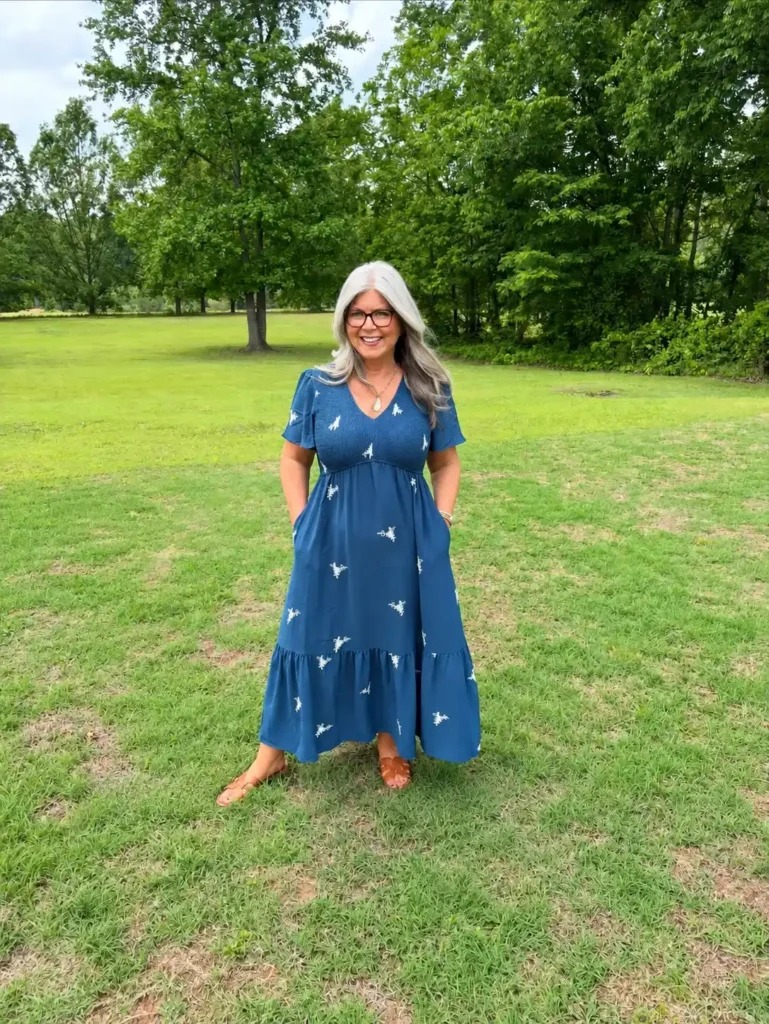Petite plus-size woman with long flowing silver-grey hair and clear-frame glasses, smiling broadly with hands relaxed at her sides, wearing a steel blue short-sleeve flutter-sleeve V-neck maxi dress with a smocked empire bodice in a honeycomb gather pattern, a full tiered A-line maxi skirt, and scattered small white botanical-style motifs across the fabric, paired with tan flat huarache-style open sandals and a simple stone pendant necklace, standing on green grass in an open park setting with large leafy trees in the background under a cloudy summer sky.