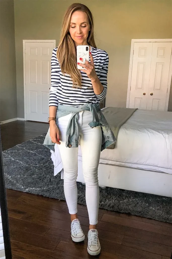 Petite woman with long straight brown hair taking a bedroom mirror selfie, wearing white slim-fit ankle-length skinny jeans, a navy and white horizontal stripe long-sleeve fitted top, a light-wash denim jacket tied around the waist by its sleeves, and white low-top Converse canvas sneakers, standing in a bedroom with a grey shag rug, white bed, and dark wood floors visible in the mirror reflection.