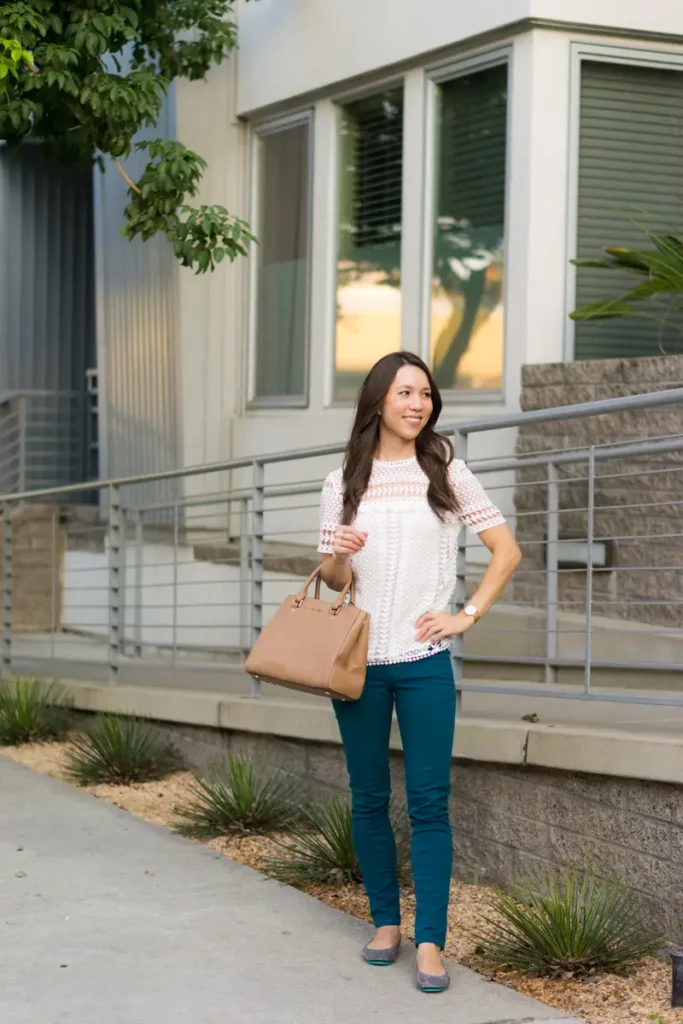 Petite woman standing on an outdoor sidewalk wearing a white short-sleeve lace crochet top with decorative floral and geometric detail at the yoke, deep teal slim-fit skinny jeans with a slight ankle crop, grey ballet flats, and carrying a tan structured satchel with gold hardware, with one hand on her hip, dark hair worn down, smiling slightly off to the side.