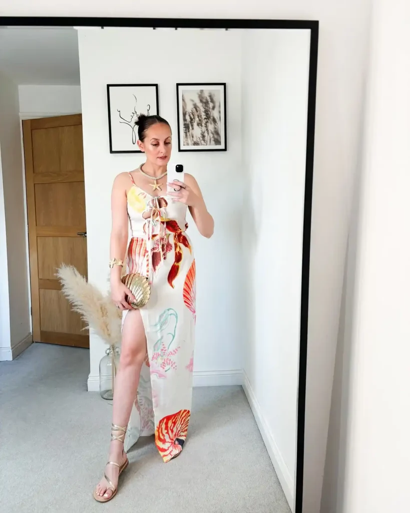 Petite woman in a cream ivory spaghetti strap maxi dress with a large-scale tropical floral and shell print in red, yellow, pink and blue, featuring a lace-up front bodice detail and a high thigh slit, paired with gold flat lace-up strappy sandals, a gold shell-shaped clutch, a gold starfish pendant layered necklace and a gold cuff bracelet, taking a mirror selfie in a neutral bedroom.