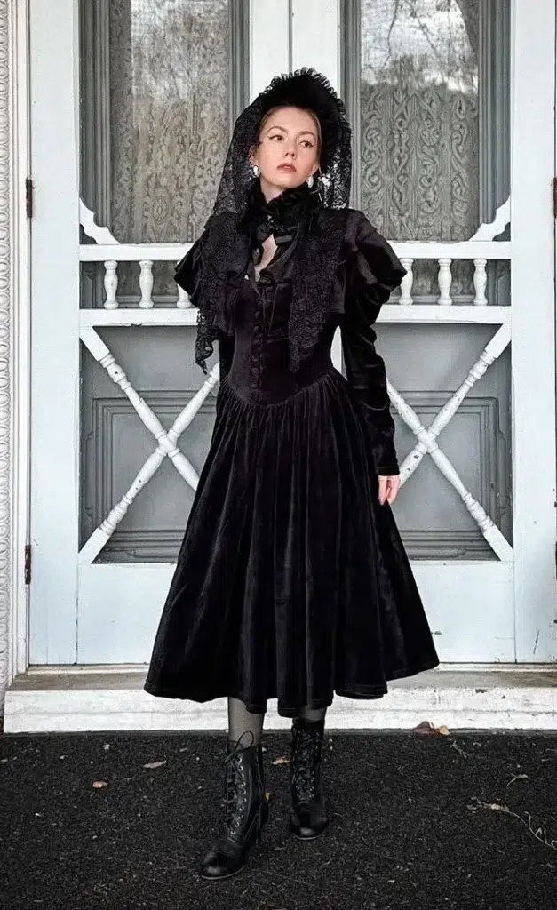 Petite woman with light hair wearing a full Victorian Gothic Halloween costume consisting of a black velvet midi dress with a fitted structured bodice, puffed long sleeves, and a sweeping full A-line skirt, layered with a black lace ruffled fichu collar piece draped over the shoulders, a black lace mantilla veil on the head, and black lace-up Victorian ankle boots with dark sheer stockings, standing before a pale blue vintage door with ornate trim.