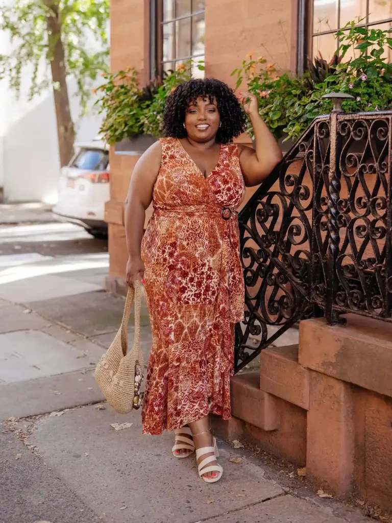 Petite plus-size woman with voluminous natural dark curly hair and small gold hoop earrings, smiling broadly with one hand raised near her head, wearing a sleeveless V-neck surplice-wrap midi dress in a dense abstract paisley-adjacent print of rust, burnt cream, and deep burgundy with a side ring-buckle waist detail and a gently flared skirt with a soft flounce near the hem, carrying a large natural woven crochet hobo bag with wooden bead handles, wearing nude multi-strap block heel sandals with an ankle strap, standing on a sidewalk next to ornate black wrought iron railings and terracotta brownstone steps with lush green trailing foliage in the background.