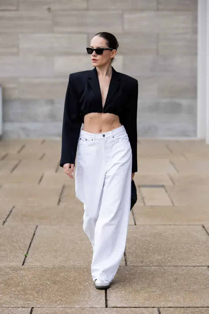 Woman with dark hair slicked back in a tight bun wearing white ultra-wide-leg low-rise baggy denim jeans with a floor-grazing hem, a cropped structured black blazer with wide lapels and strong shoulder seams ending just below the ribcage, and white low-profile sneakers, with black narrow cat-eye sunglasses, walking on an outdoor stone pavement in front of a grey stone wall.