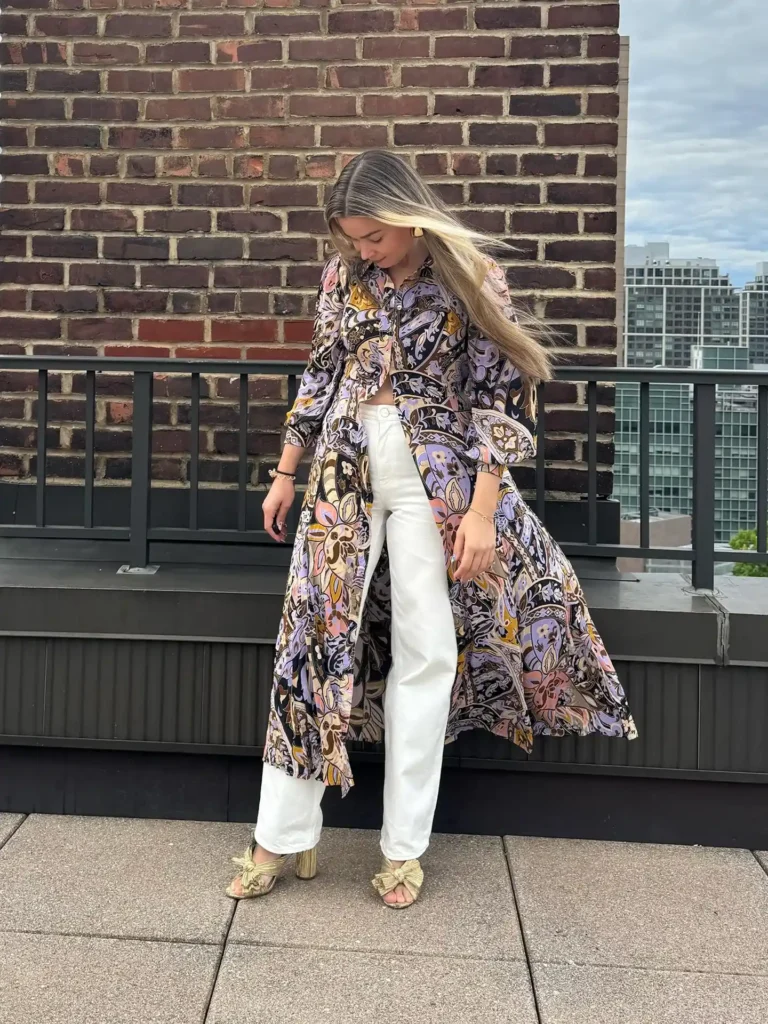Woman with long blonde hair standing on an urban rooftop terrace, wearing white high-rise wide-leg denim jeans with a visible button waistband, a floor-length open printed maxi shirt dress with a bold large-scale psychedelic paisley pattern in lavender, black, mustard gold, brown, and blush pink worn fully unbuttoned as a duster layer, and gold metallic bow-tie heeled mule sandals, with gold drop earrings, with a red brick building wall and city skyline visible in the background.