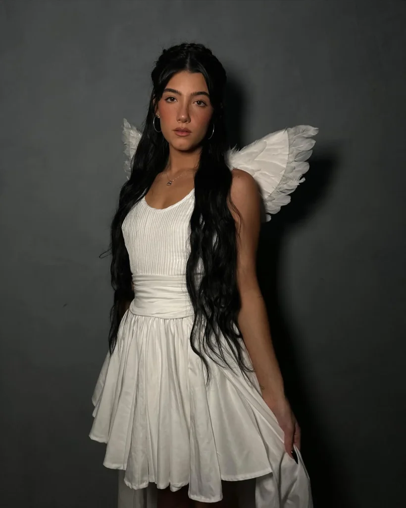 Petite woman with long dark wavy hair wearing a white Halloween angel costume consisting of a white ribbed fitted bodice, wide white wrapped waist sash, and a full white flared mini skirt, styled with white feather angel wings attached at the back, photographed against a dark gray studio background.