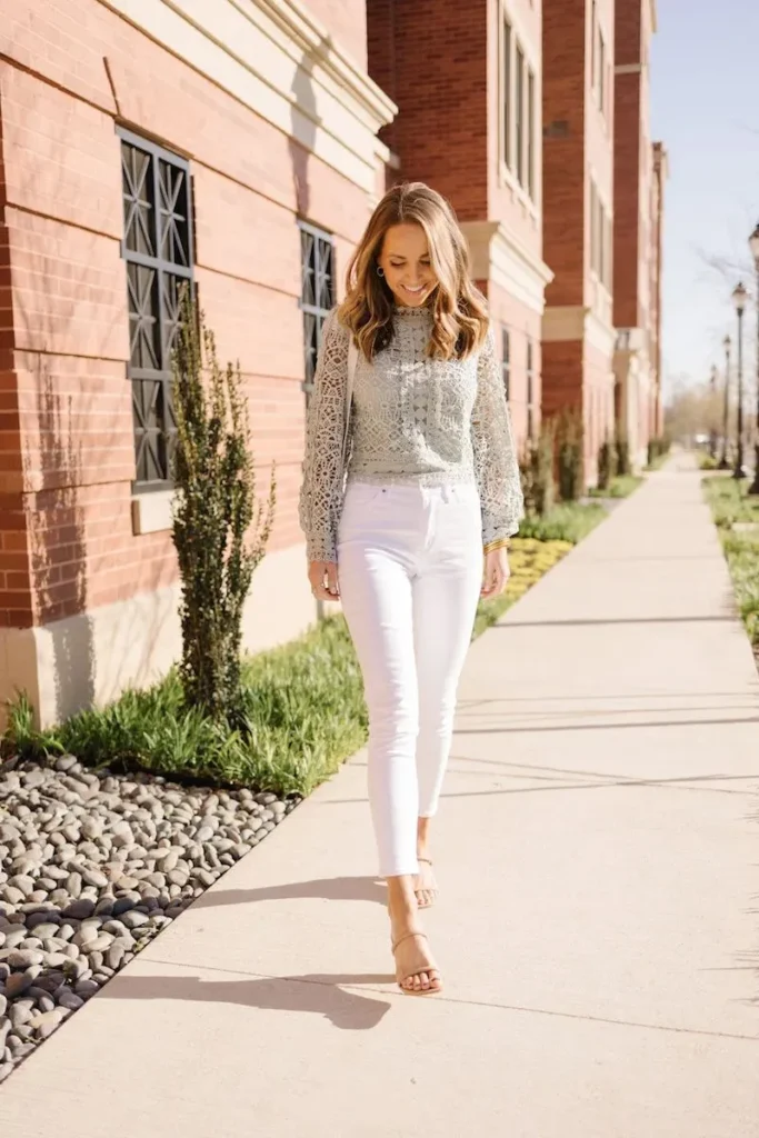 Petite woman in white slim-fit high-rise ankle-crop jeans and a sage mint green crochet lace high mock-neck long-sleeve balloon-sleeve top, wearing nude strappy heeled sandals, walking and smiling on an outdoor sidewalk beside a red brick building on a bright sunny spring day.
