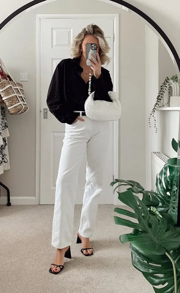 Woman with short wavy blonde hair taking a mirror selfie inside a round arched black-framed mirror, wearing white high-rise straight-leg full-length denim jeans, an oversized textured black button-up shirt loosely front-tucked with a thin white belt, black strappy block-heeled mule sandals, a small white woven intrecciato-style mini clutch bag carried with a pearl chain strap, and a pearl chain necklace, standing in a neutral-toned bedroom with a large monstera plant beside the mirror and a woven basket bag hanging on the left.
