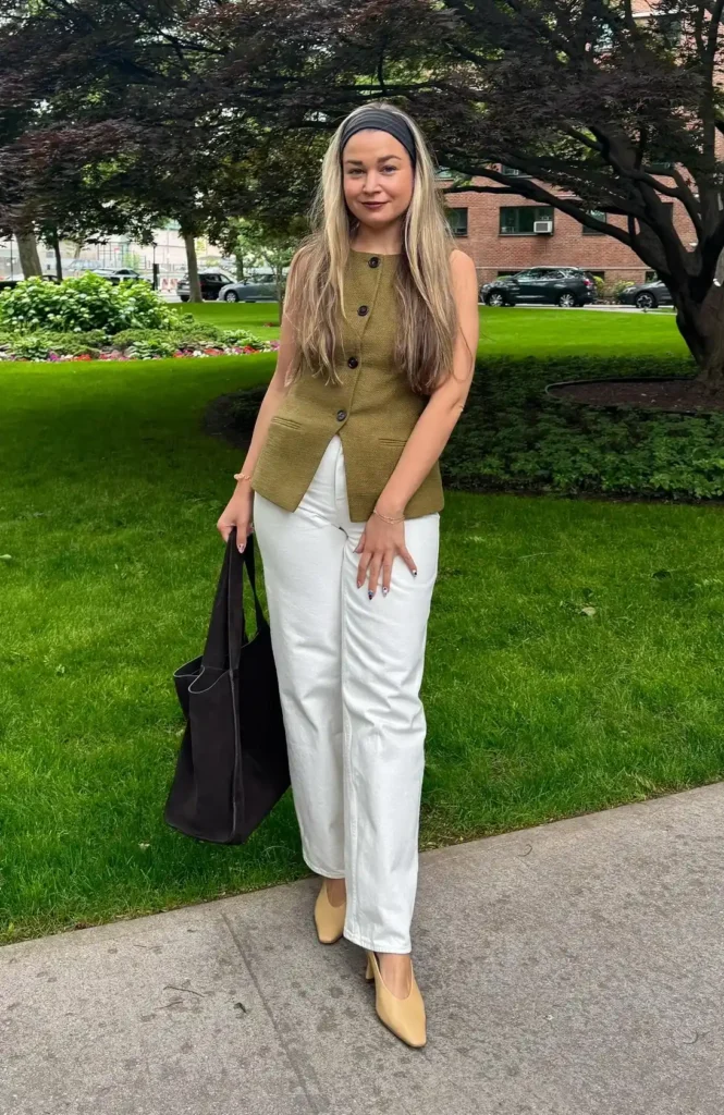 Petite woman with long blonde hair wearing a dark grey fabric headband, dressed in white high-rise wide-leg denim jeans with a clean ankle hem, a fitted olive green textured sleeveless button-front waistcoat vest with dark buttons, butter yellow kitten heel square-toe pumps, and carrying a large soft black tote bag with top handles, standing on a sidewalk path beside a lush green park lawn with a burgundy Japanese maple tree and red brick buildings visible in the background.