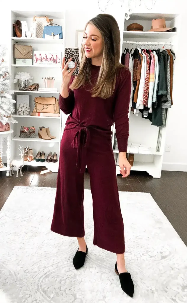 Petite woman taking a mirror selfie in a matching deep wine knit set consisting of a long-sleeve crewneck top with a fabric self-tie side knot at the waist and wide-leg culotte-length pants in the same color, paired with black pointed-toe slip-on mules, standing on a white rug in front of a white shelving unit displaying handbags and shoes, with a clothing rack visible in the background.