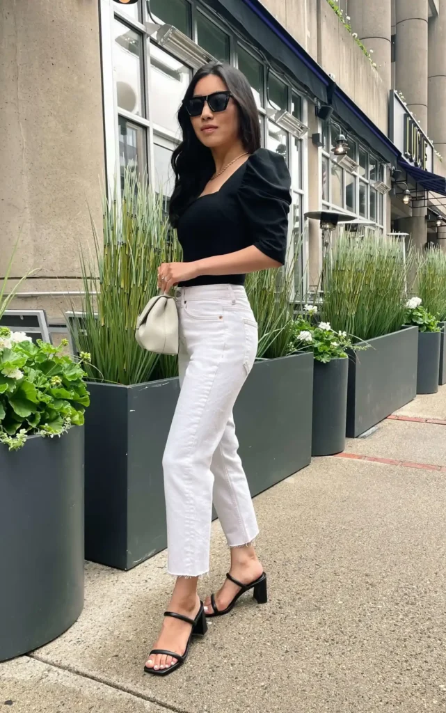 Petite woman with long dark hair wearing white high-rise straight-leg cropped jeans with a raw frayed hem, a fitted black square-neck short puff-sleeve top, black double-strap block-heeled sandals, and a small soft cream slouchy bag held at the wrist, with a gold chain necklace and black cat-eye sunglasses, standing on an outdoor urban sidewalk beside tall dark planters with ornamental grasses.
